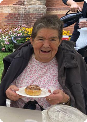 Elderly woman smiling and holding a plate with a pastry while sitting outdoors near flowers and a brick wall.