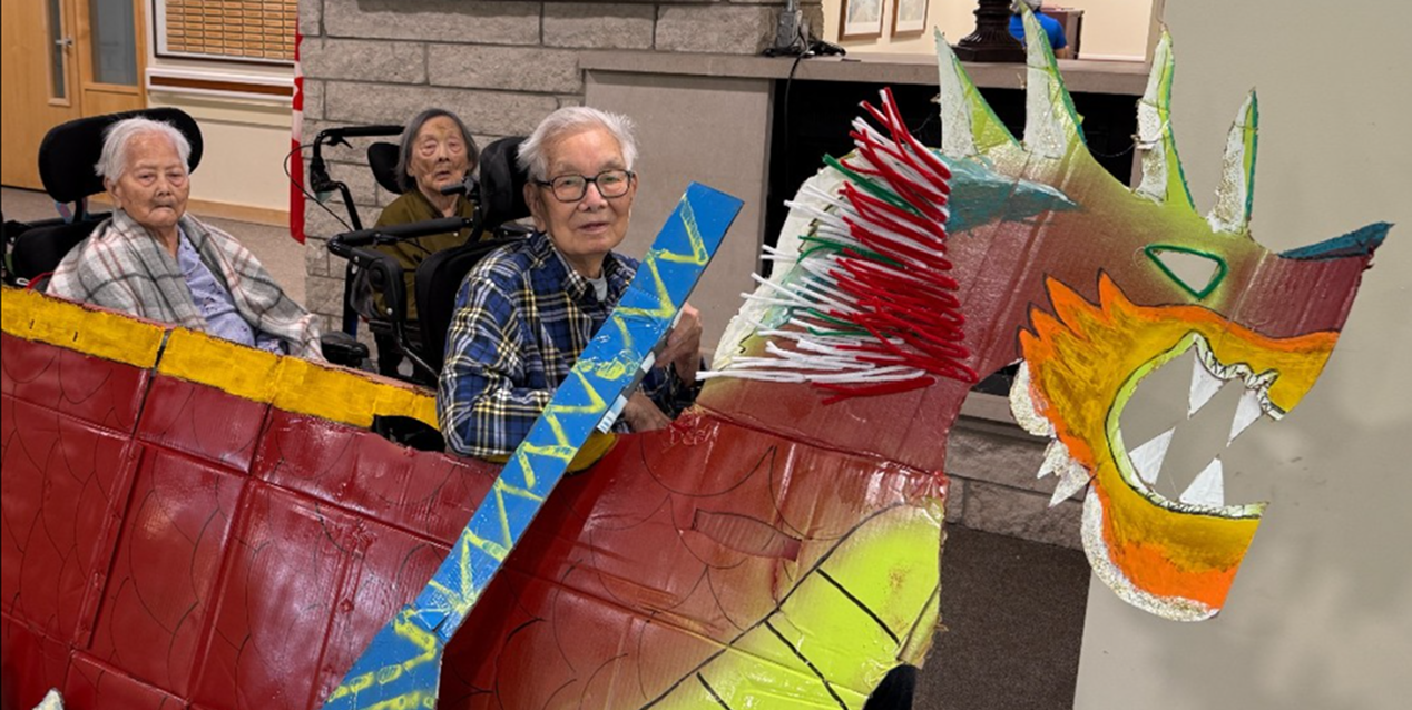 Three elderly people seated in wheelchairs participate in a colorful, handmade dragon boat craft event indoors.