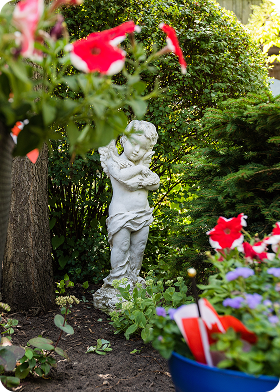 White cherub statue holding a bird in a garden surrounded by green bushes and colorful flowers.
