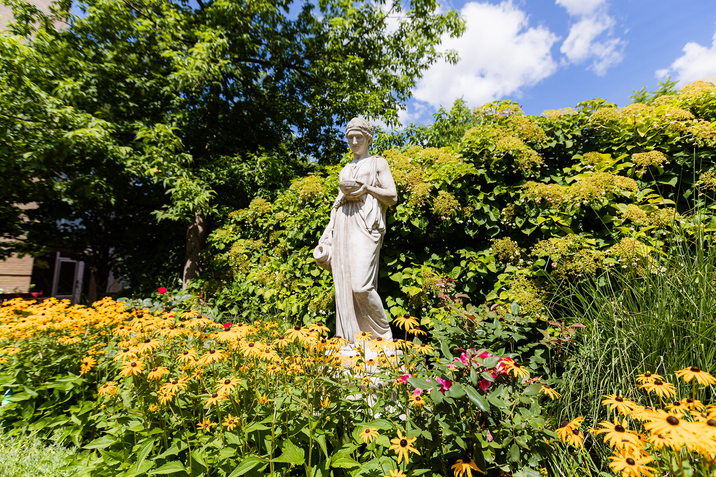 White classical statue of a woman holding a bowl and jug surrounded by yellow flowers and green bushes under a blue sky with clouds.