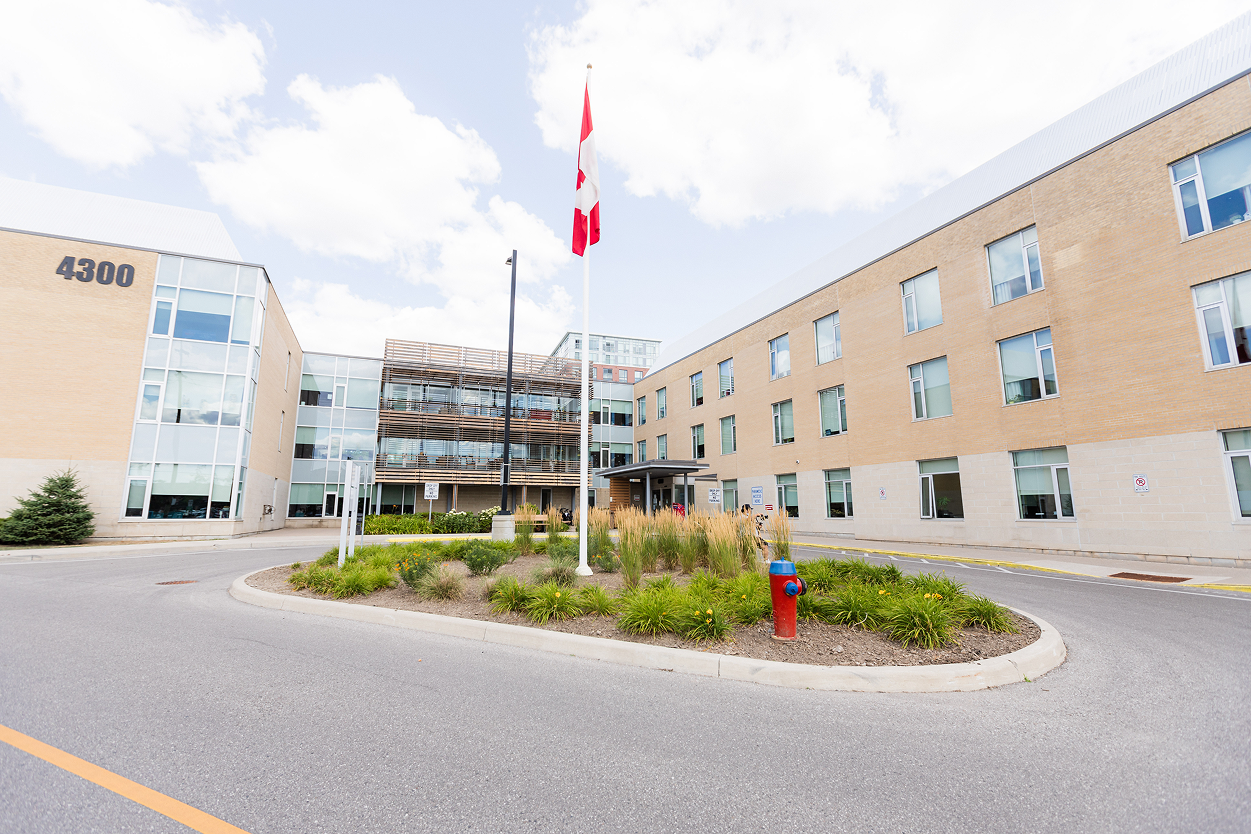 Modern hospital building with a Canadian flag on a pole and a circular garden with bushes and a fire hydrant in front.