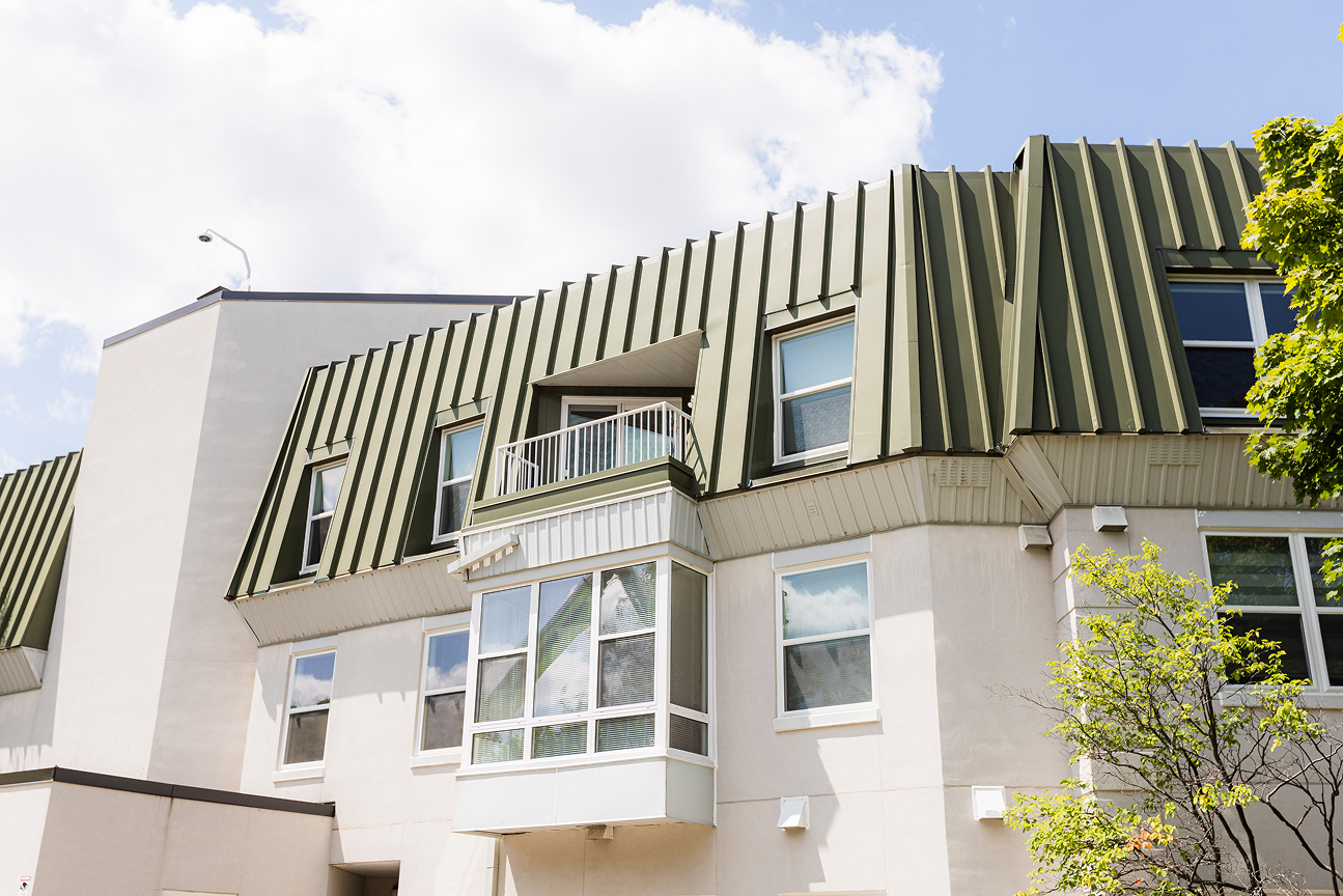 Modern residential building with green metal mansard roof, white walls, multiple windows, and a small balcony under blue sky with clouds.