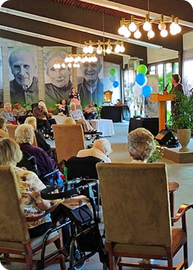 Group of elderly people seated in a room listening to a speaker at a podium, with large portraits on the wall and colorful balloons nearby.