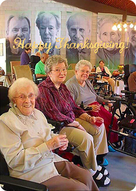 Three elderly women smiling and seated in a communal room with 'Happy Thanksgiving' and large portraits of older men displayed on the wall behind them.