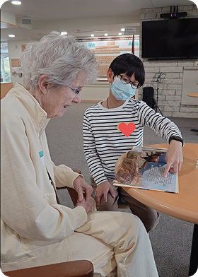 A young person wearing glasses and a face mask reads a book to an elderly woman in a bright room.