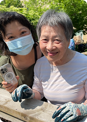 Smiling elderly woman wearing gardening gloves sitting at a wooden table outdoors with a younger woman beside her wearing a face mask and holding a smartphone.