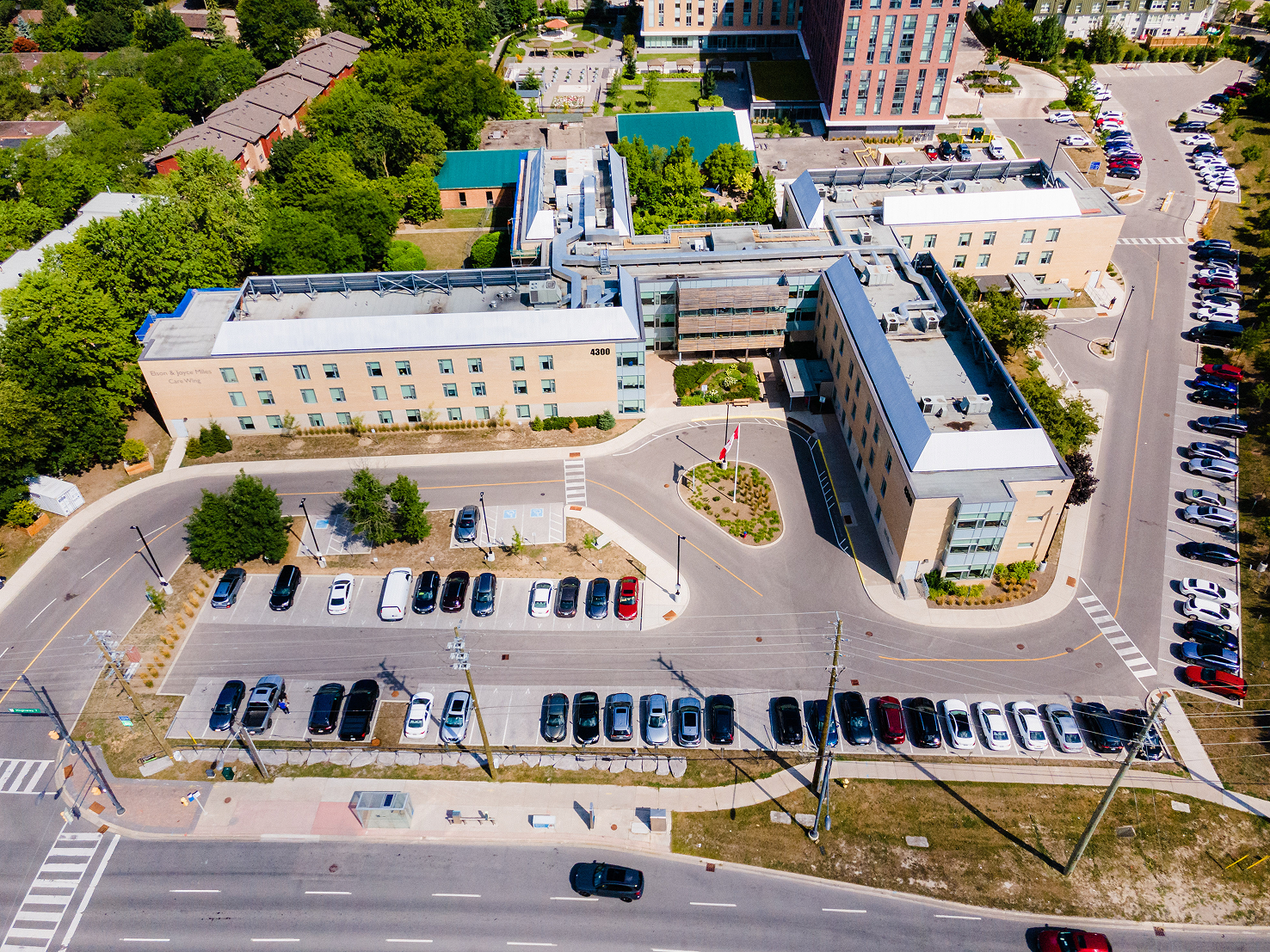 Aerial view of a multi-wing building complex with parking lots, a Canadian flag, and surrounding trees and roads.