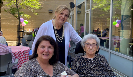 Three women smiling outdoors at a table with pink, green, and purple balloons in the background.