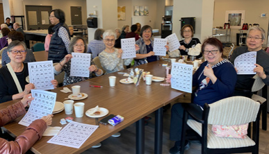 Group of elderly women sitting around a table, smiling and holding up bingo cards in a community room.