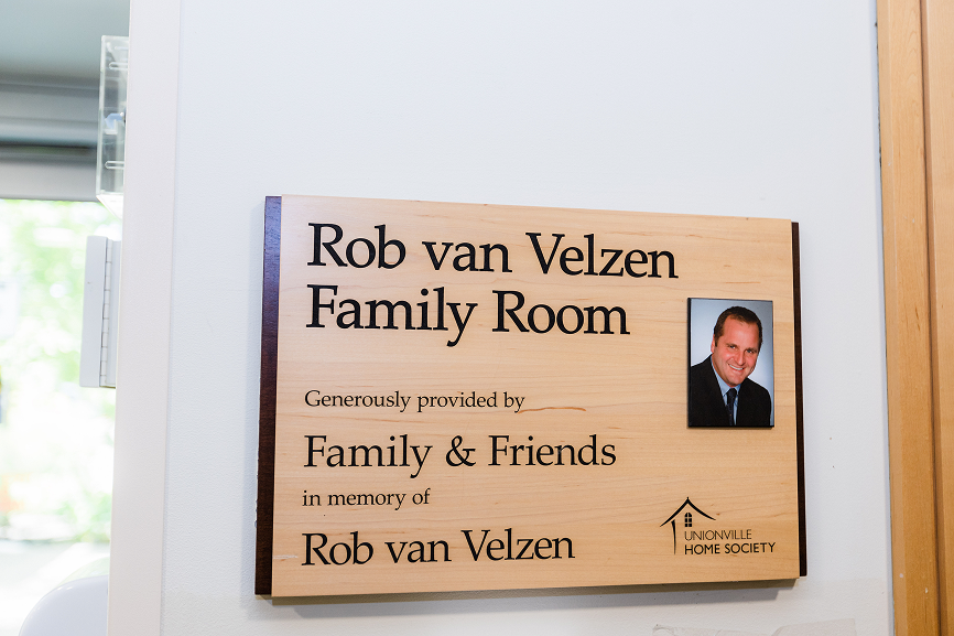 Wooden plaque on a wall reading 'Rob van Velzen Family Room, Generously provided by Family & Friends in memory of Rob van Velzen' with a photo of a smiling man in a suit.