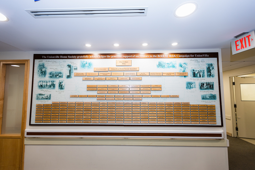 A large donor recognition plaque with multiple engraved name plates mounted on a wall under ceiling lights, with an EXIT sign and doors nearby.