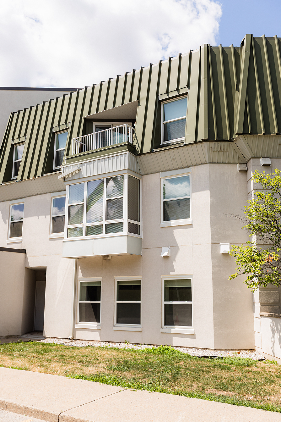 Exterior view of a beige multi-story building with olive green metal mansard roof and several windows, partly shaded by sunlight and a small tree on the right.