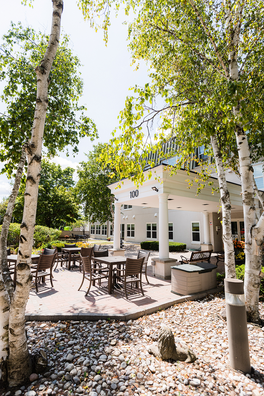 Outdoor patio with tables and chairs surrounded by birch trees in front of a building entrance numbered 100.