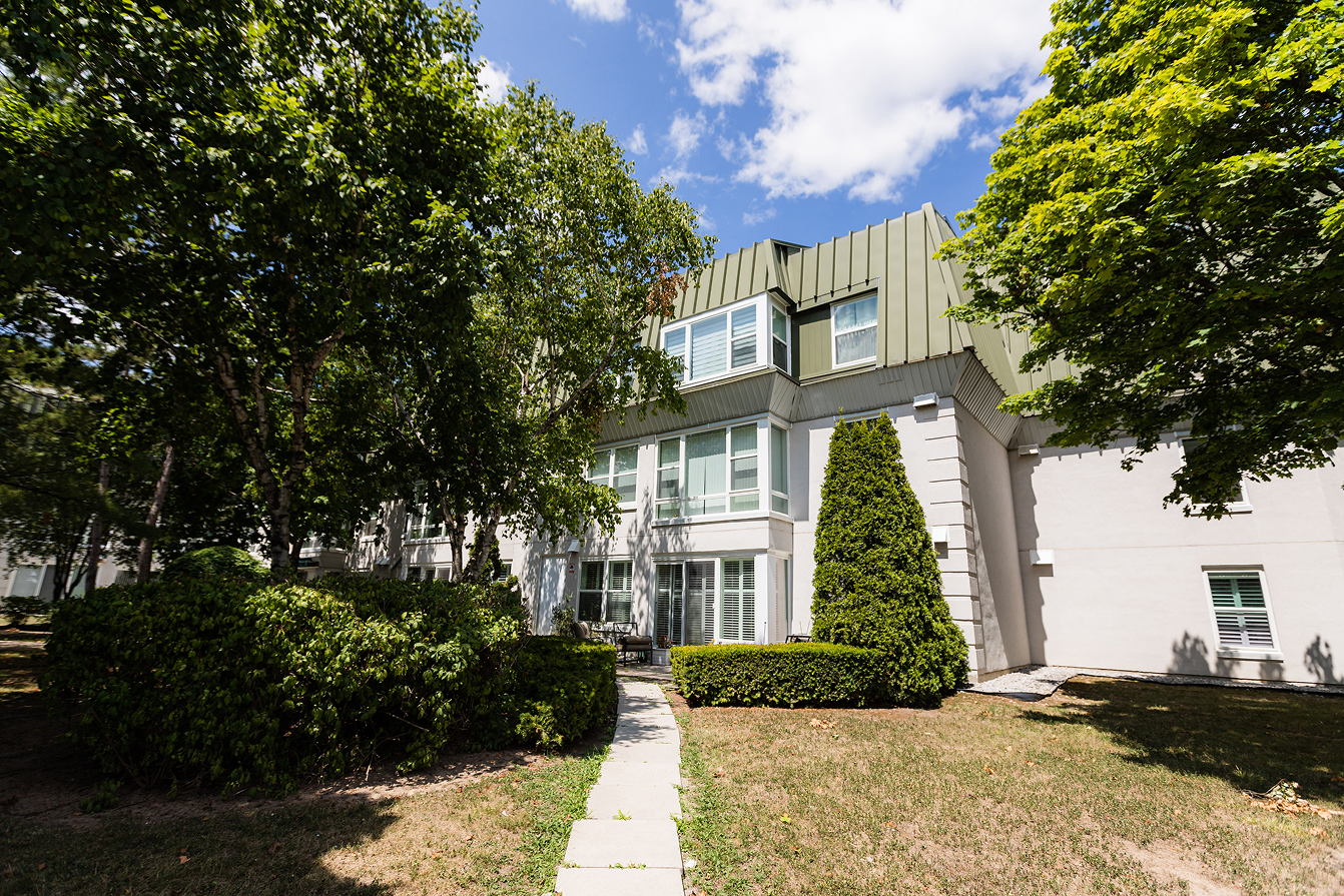 Three-story residential building with green roof and large windows surrounded by green trees and bushes under a blue sky with scattered clouds.