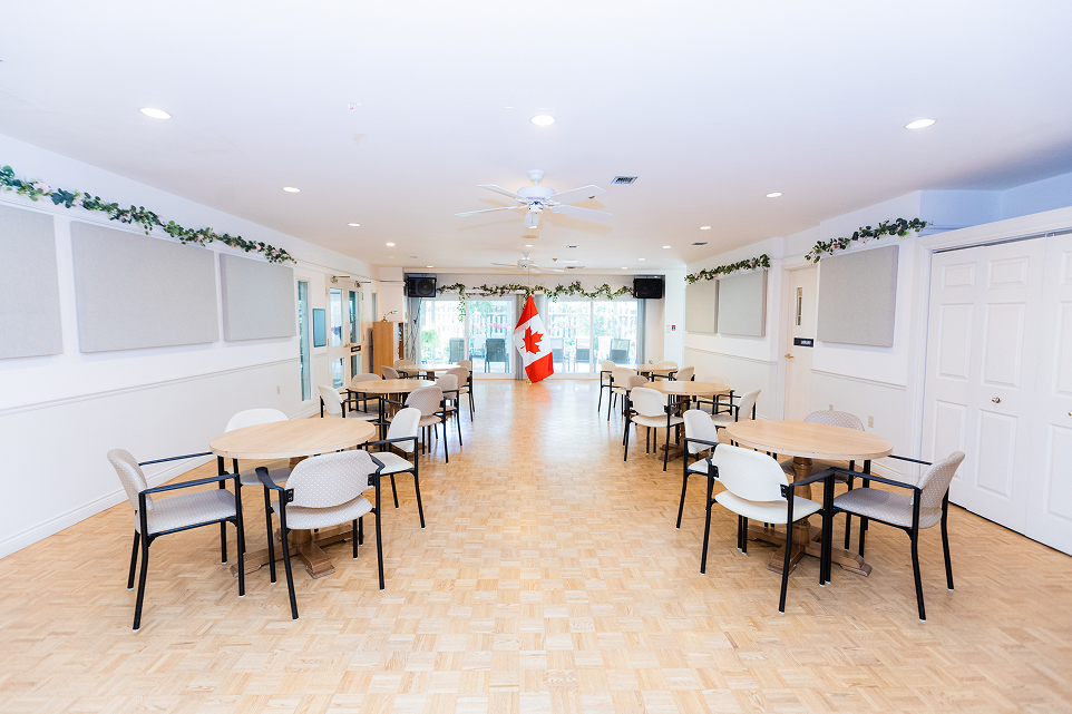 Bright community room with round tables and chairs, decorated with greenery and a Canadian flag at the far end.