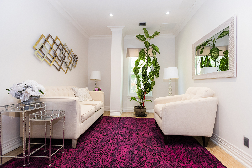 Bright living room with two beige upholstered sofas facing each other across a deep purple patterned rug, a tall green plant near a window, mirrored wall decor, and two table lamps.