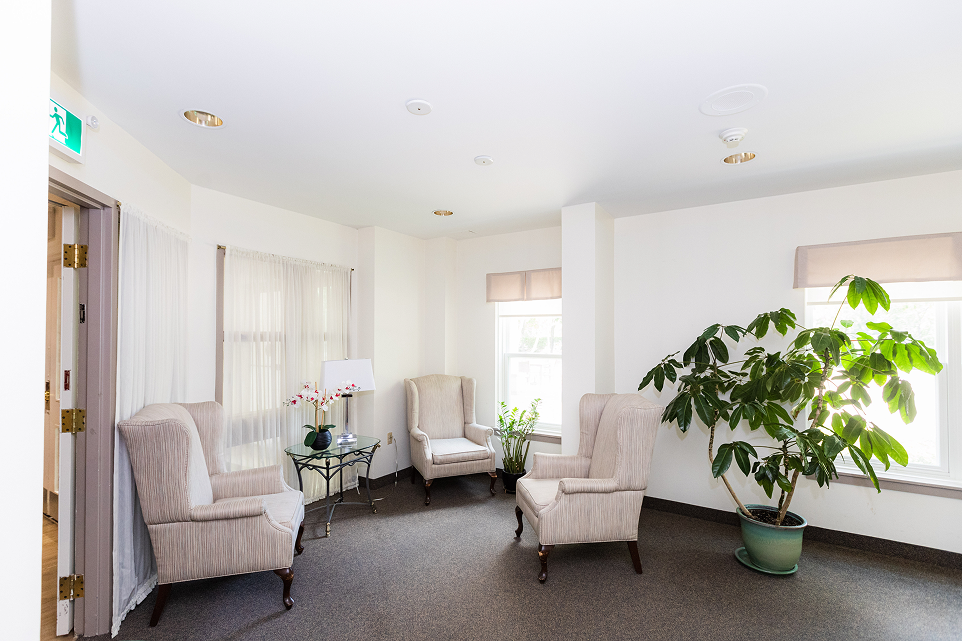Bright sitting area with three beige armchairs arranged around a glass-top side table holding a lamp and an orchid, with two large potted plants near the windows.
