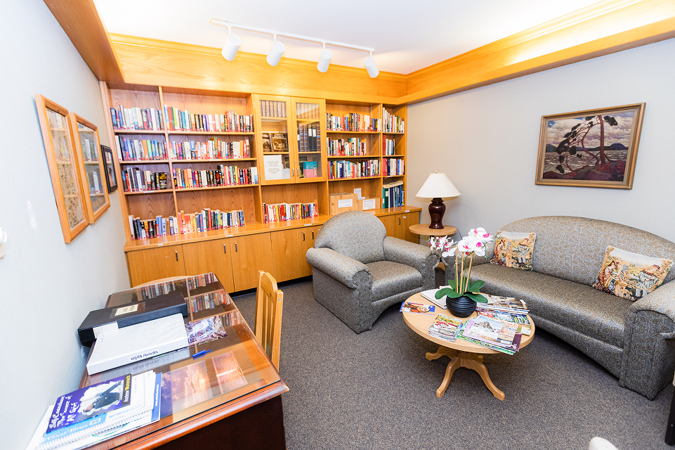 Cozy reading room with wooden bookshelves filled with books, a gray armchair, a matching couch with decorative pillows, a round wooden coffee table with magazines and a vase of white orchids, a side table with a lamp, and framed artwork on the wall.