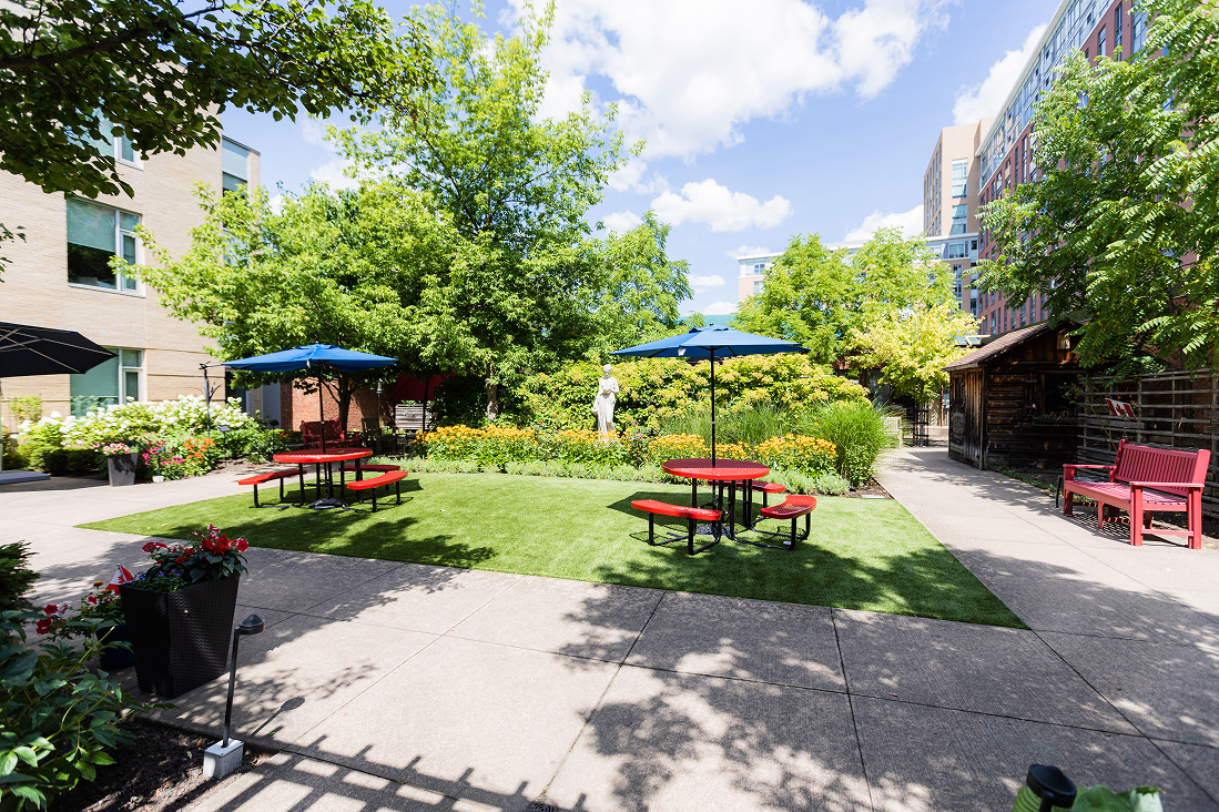 Outdoor courtyard with green grass, two red picnic tables under blue umbrellas, surrounded by trees, flowers, and buildings.