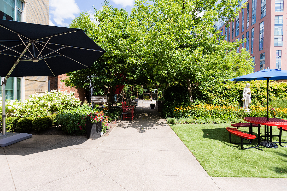 Outdoor courtyard with green trees and bushes, red picnic tables with blue umbrellas, benches, flowers, and a statue near modern buildings.