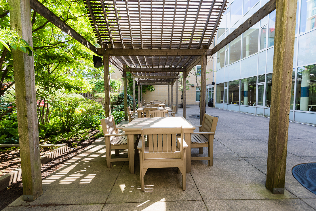 Outdoor wooden tables and chairs under a pergola next to a modern building with large windows and surrounding greenery.