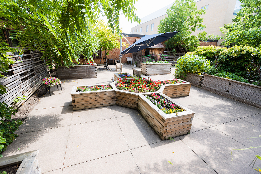 Outdoor patio garden with wooden raised flower beds arranged in geometric shapes, colorful flowers, green trees, and a large dark umbrella.