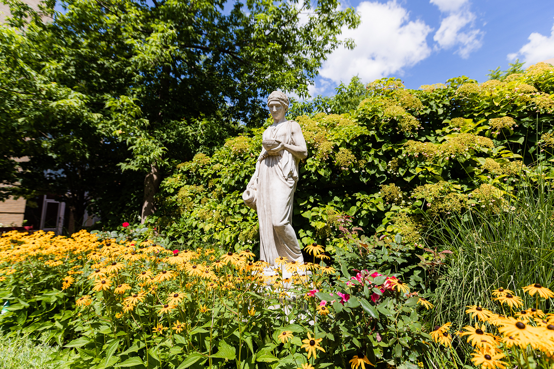 White stone statue of a woman holding a bowl and pitcher surrounded by yellow and pink flowers and lush green foliage under a blue sky.
