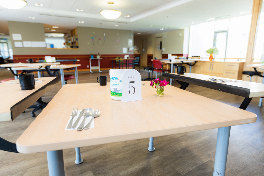 Empty cafeteria with wooden tables, one table labeled with a card showing number 5, cutlery on a napkin, a small flower vase, and a container of wipes.