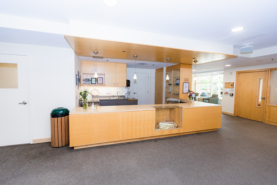 Modern reception desk with light wood finish, pendant lights, flower vases, and a small decorative Christmas tree inside a glass display.