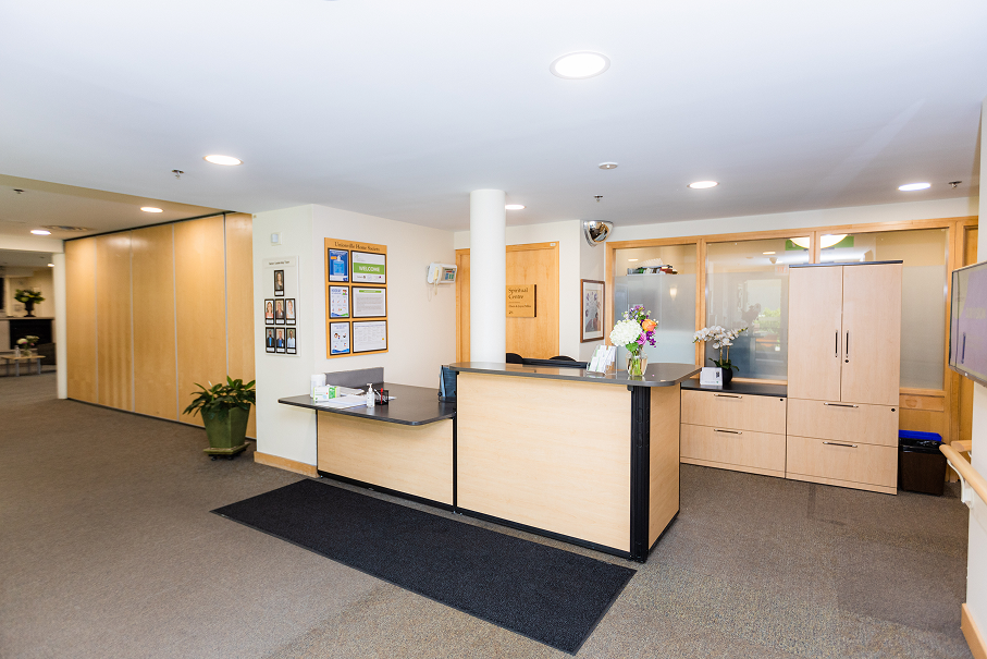 Reception desk in a clean, well-lit office with wooden cabinetry, informational posters on the wall, and potted plants.