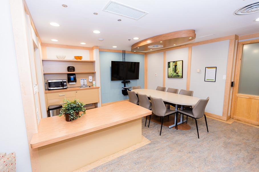 Modern conference room with a long oval table, eight chairs, a wall-mounted flat screen TV, a wooden countertop with a plant, and a kitchenette with a microwave and coffee machine.