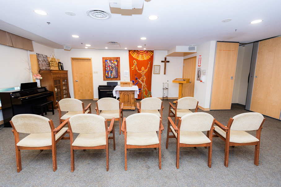 Small chapel room with two rows of beige cushioned wooden chairs facing a wooden pulpit, decorated with a red tapestry, a cross, and paintings on the wall.
