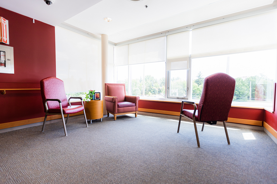 Bright room with large windows, three red cushioned chairs, a small round wooden table with a plant and framed photo, and maroon accent walls.