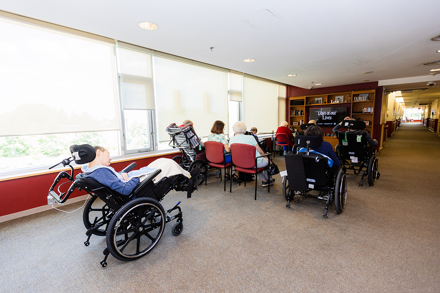 Group of elderly individuals, some in wheelchairs, seated in a common room watching a television screen displaying 'Days of our Lives.'