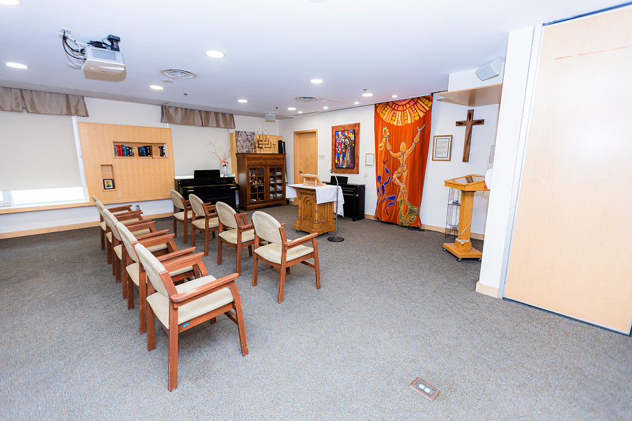 Small chapel with wooden chairs arranged in two rows facing a lectern and a colorful religious tapestry on the wall.