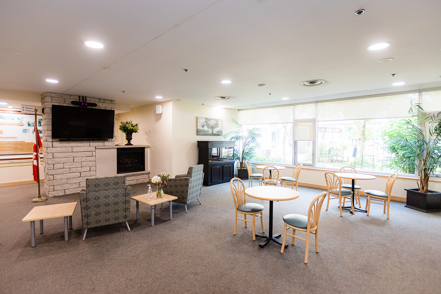 Bright lounge area with round tables and wooden chairs, two patterned armchairs facing a TV above a stone fireplace, large windows with greenery outside, and potted plants indoors.