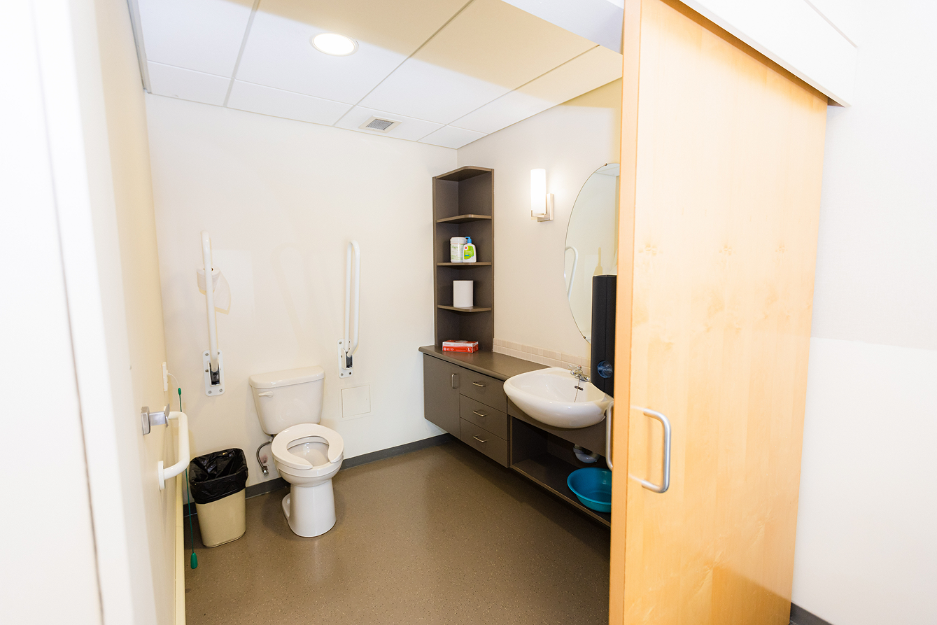 Accessible restroom with white toilet, grab bars, a trash bin, a sink, an oval mirror, shelves with supplies, and a sliding wooden door.