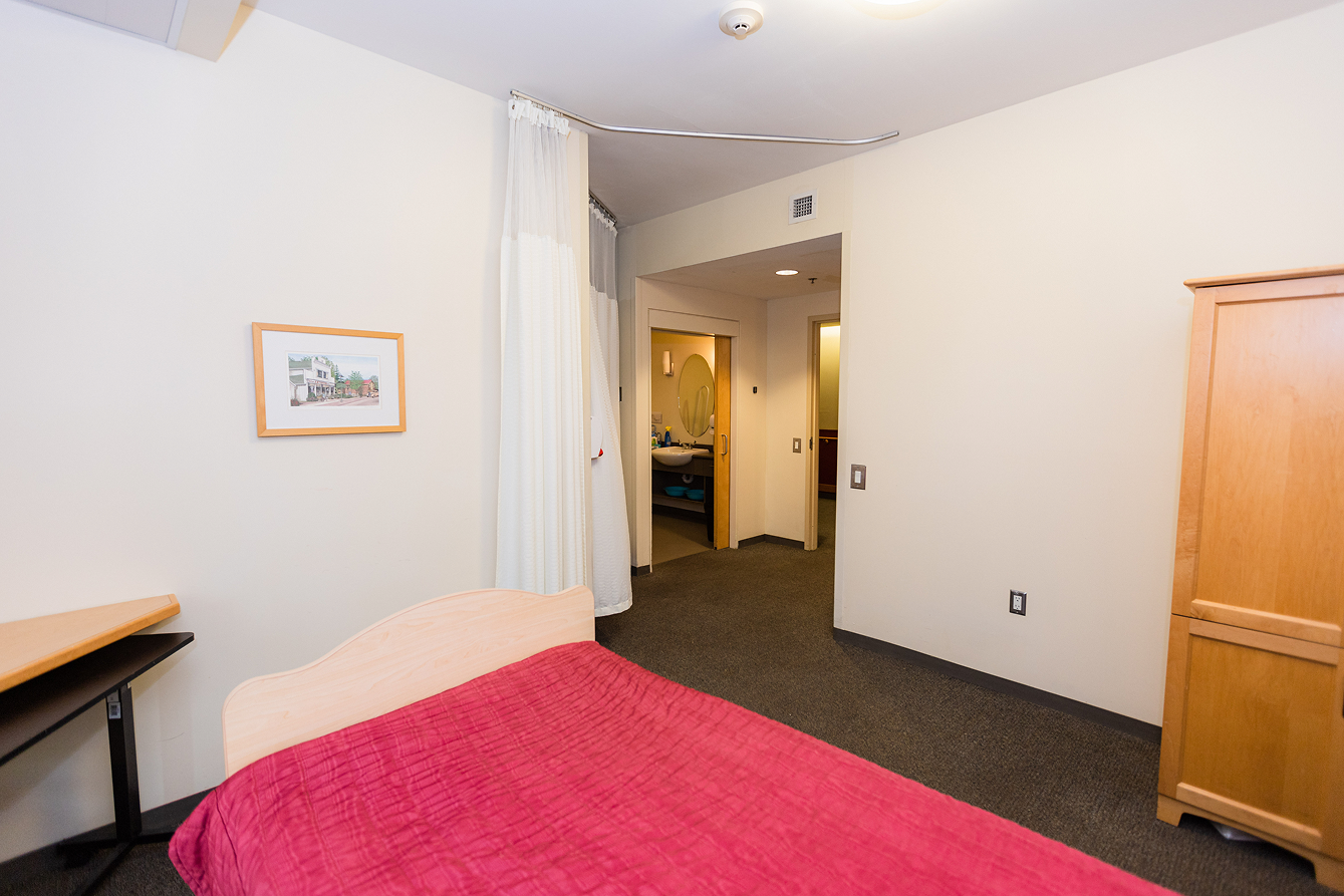 Simple room with a bed covered by a red blanket, a wooden cabinet, a small desk, a framed picture on the wall, and an open doorway leading to a bathroom with a sink and mirror.