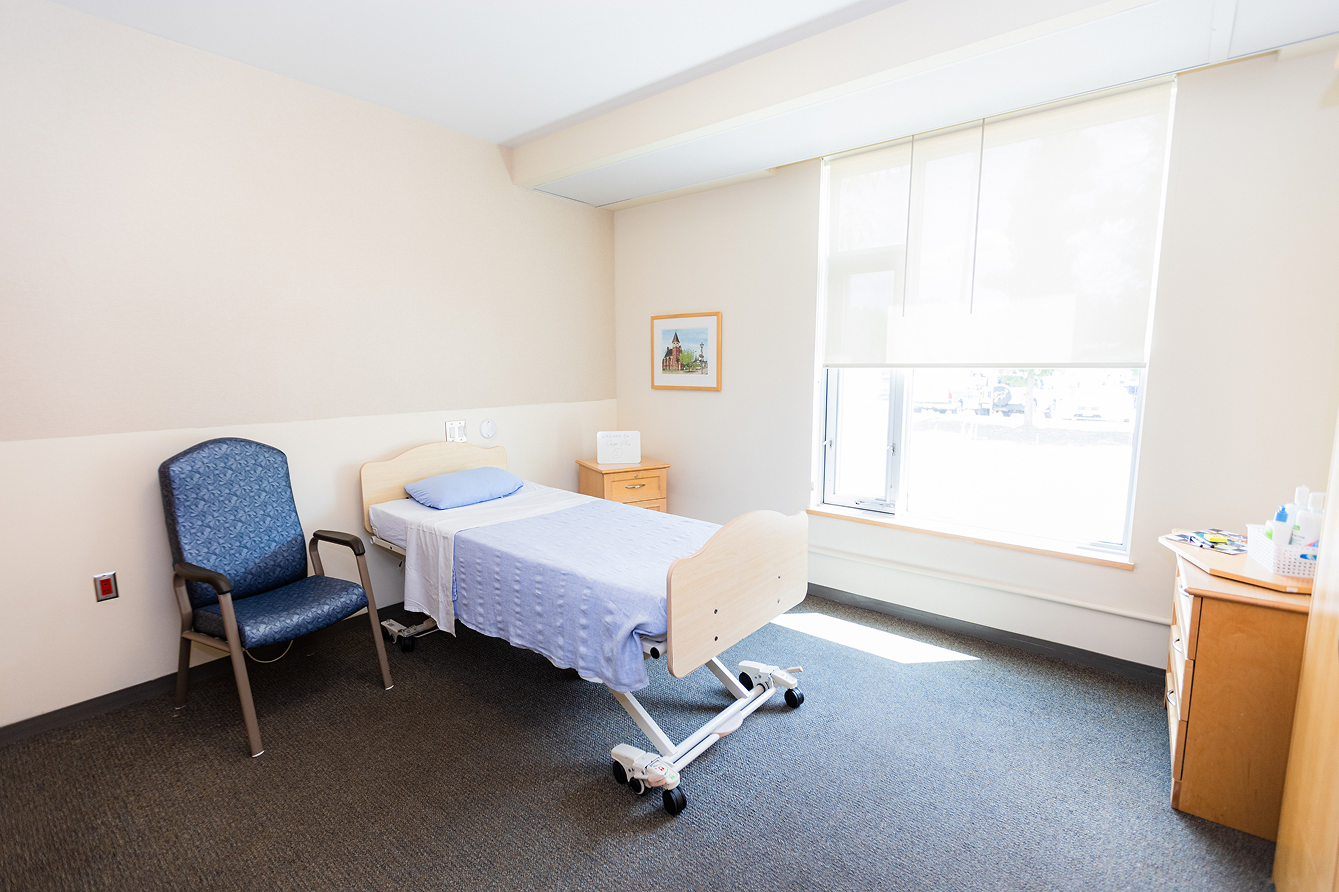 Bright hospital or care room with a single bed covered by a light blue blanket, a blue patterned chair, a window with blinds, and wooden furniture.