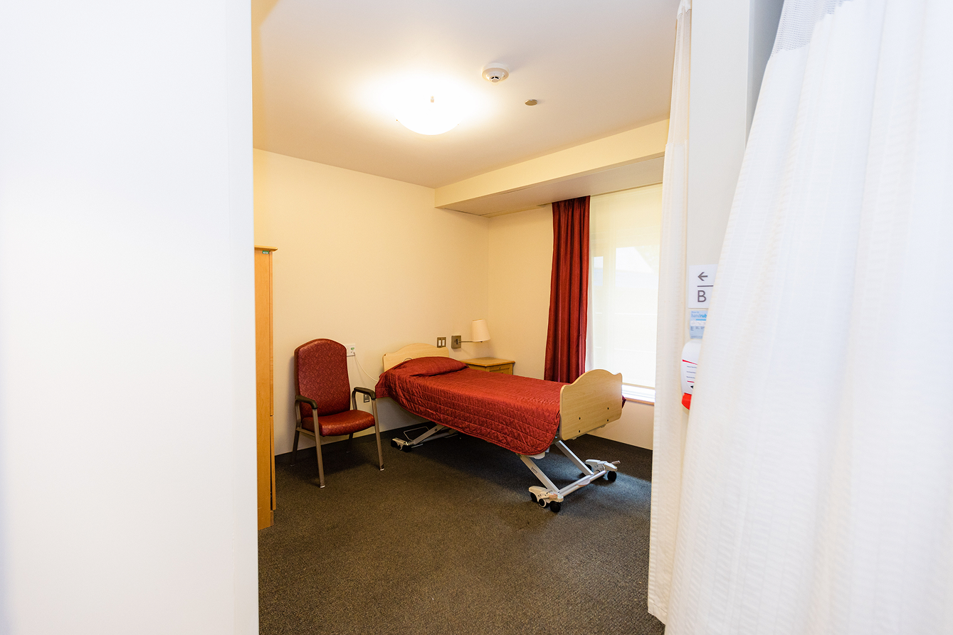 Small hospital or care facility room with a single bed covered in a red blanket, a red cushioned chair, a wooden nightstand, and a window with red curtains.