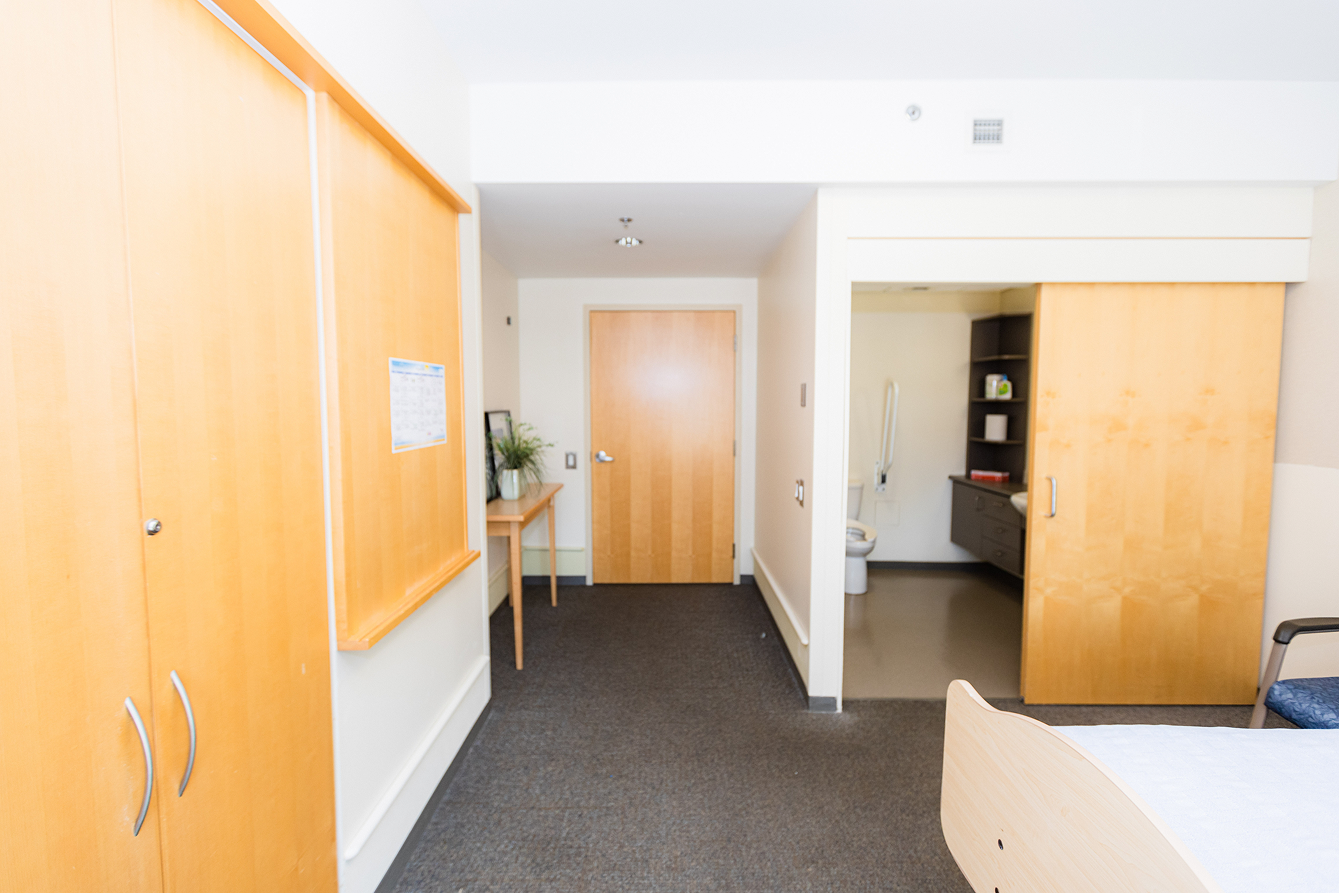 Bright hospital room with wooden furniture, a bed partially visible on the right, a closed door straight ahead, and an open sliding door showing a bathroom with a toilet and shelves.