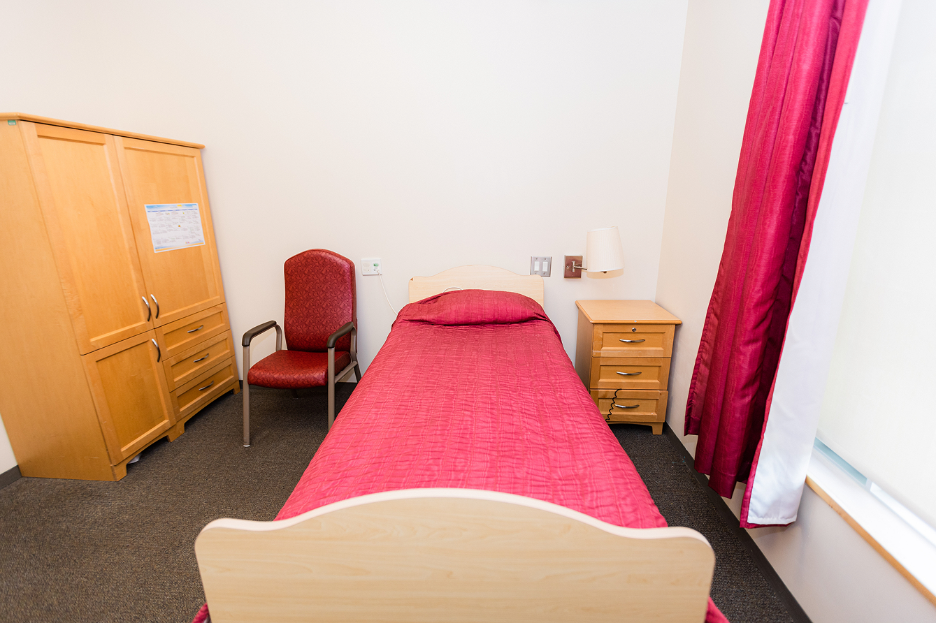 Small bedroom with a single bed covered in a red quilt, a red cushioned chair, wooden wardrobe, and bedside table with a lamp, next to a window with red curtains.