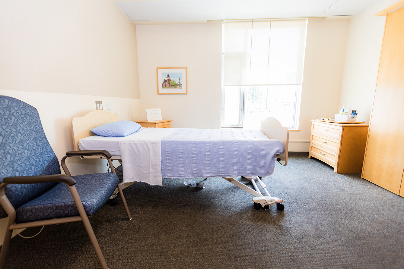 Bright hospital or care room with a single bed, blue chair, window with a roller shade, and wooden furniture.