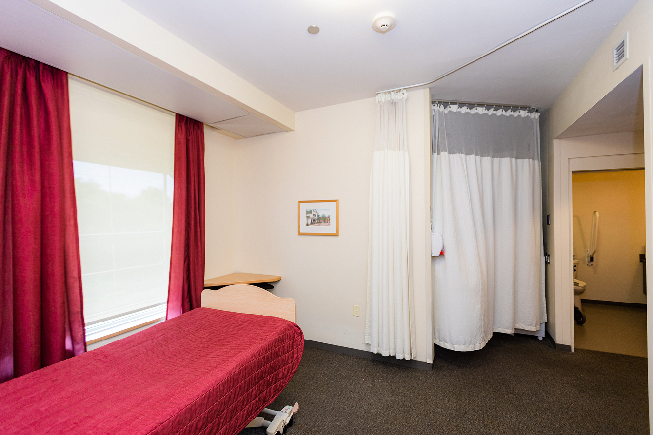 Medical examination room with a bed covered in a red blanket, red curtains on the window, a small corner desk, white privacy curtains, and an open door leading to a restroom.