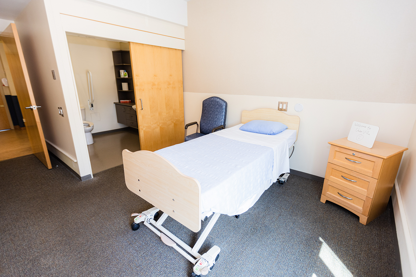 Simple hospital or care room with a single adjustable bed, a blue chair, wooden nightstand, and open bathroom door.