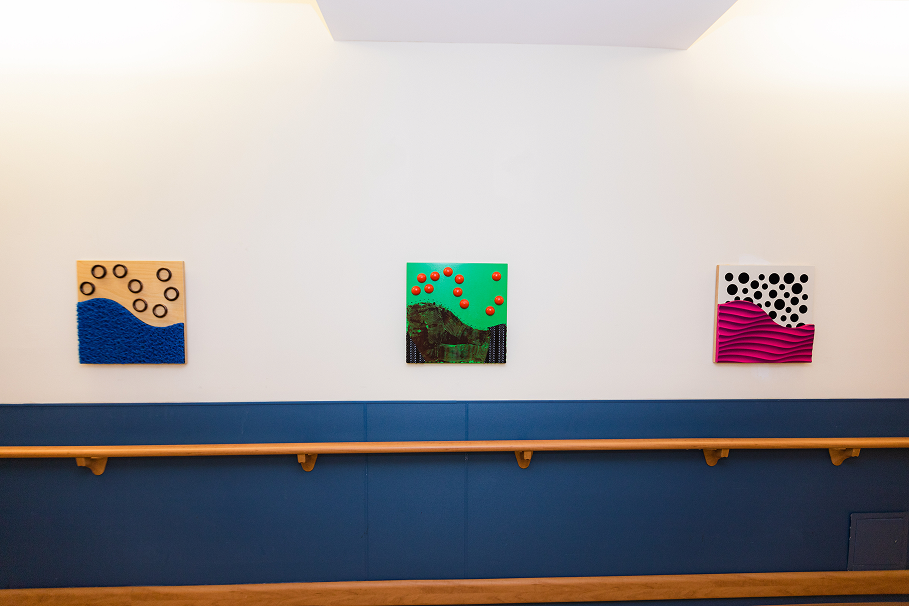 Three colorful abstract square artworks with textured waves and circular patterns mounted on a white wall above a wooden handrail and blue paneling.