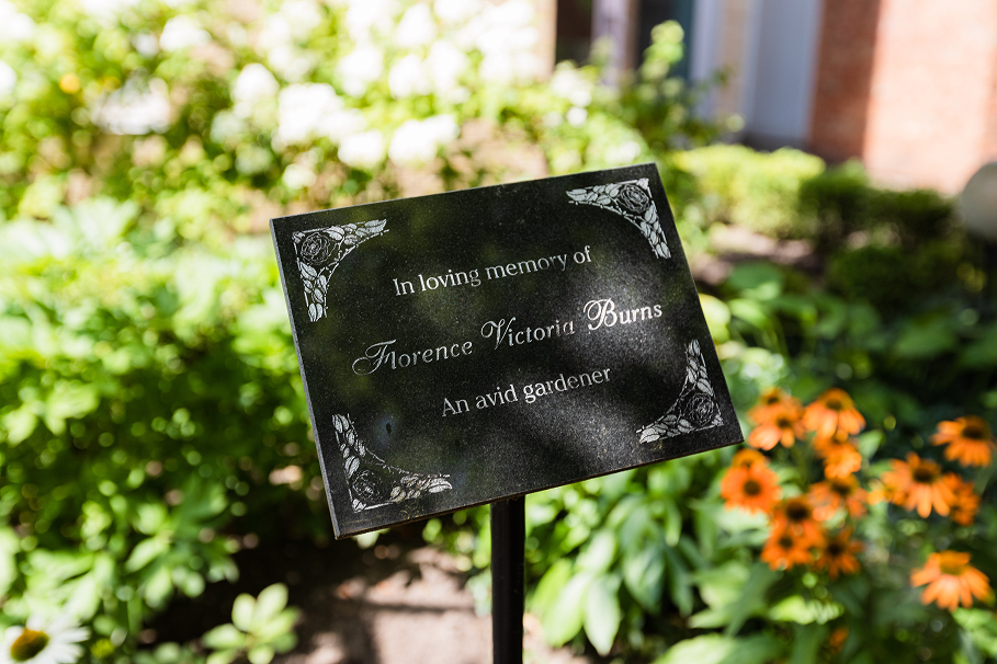 Memorial plaque reading 'In loving memory of Florence Victoria Burns, An avid gardener' surrounded by greenery and orange flowers.