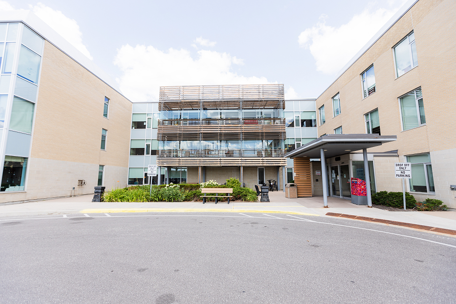 Modern three-story building with large windows and a covered entrance, surrounded by greenery and a bench in front.