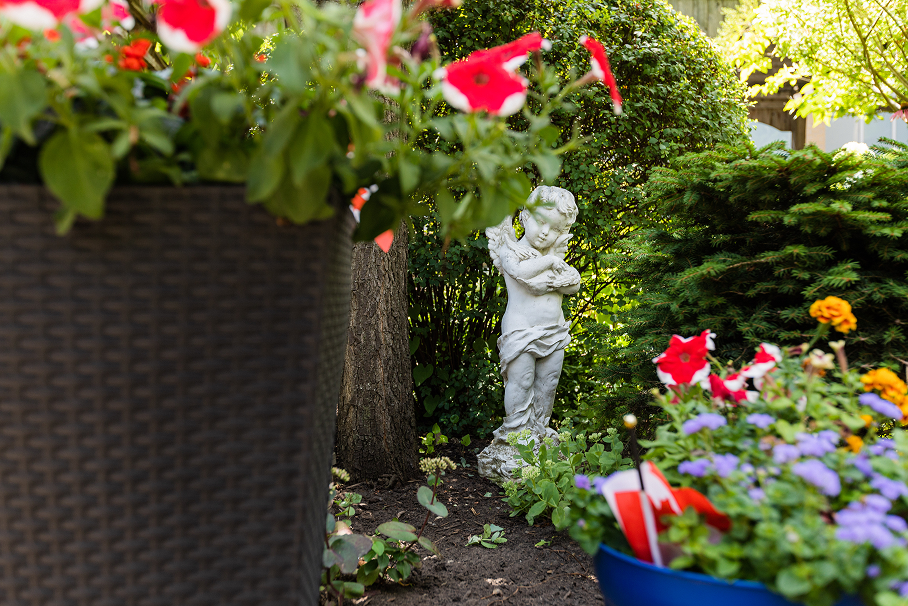 White cherub statue holding a bird surrounded by green bushes and colorful flowers in a garden.