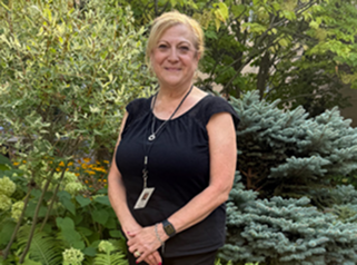 Smiling woman with short blonde hair wearing a black top standing outdoors by green and blue-green foliage.